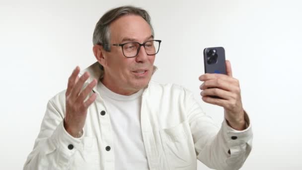 一个快乐而成熟的男人正在打一个视频电话 高兴地在智能手机的屏幕上挥手 穿着一件休闲的白色夹克 背景干净 8K照相机 — 图库视频影像