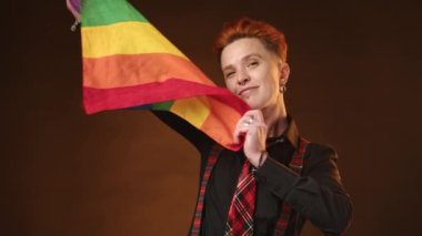 Kendine güvenen lezbiyen bir kadın, LGBT bayrağının canlı renklerini kavrıyor, sonra da kolsuz bir şekilde omzuna atıyor, karanlık bir arkaplanda gelişigüzel ama güçlü bir duruş sergiliyor. Kamera 8K HAM. 
