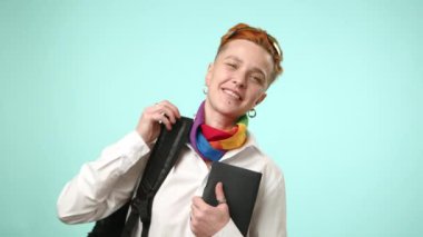 Omzuna sırt çantası takmış neşeli bir lezbiyen öğrenci kameraya sırıtıyor, LGBT eşarbı açık deniz mavisi bir zemine karşı kapsayıcılığı simgeliyor. Kamera 8K HAM. 