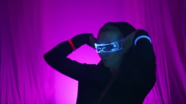 Bir sanat alanının sükunetinde holografik gözlüklü bir siber punk kız ve müziğin sakinleşmesi için kapüşonlu siyah bir pelerin sallanıyor, silueti bir ışık dansı yapıyor. Yüksek kalite 4k görüntü