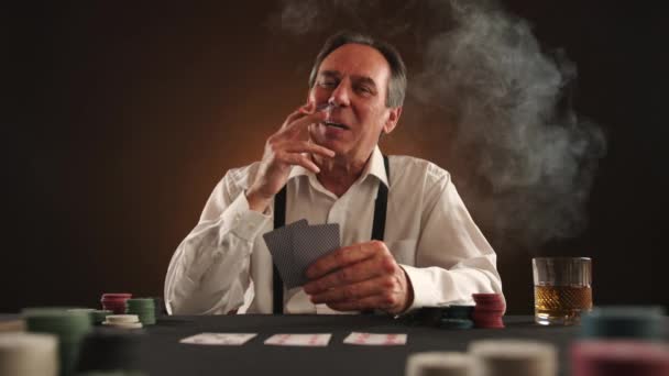 白いシャツとサスペンサーでポーカーをする成熟したビジネスマン 男はタバコを吸っている 彼の勝利に非常に自信を持って 微笑み わずかにふわふわしている テーブルの上のチップとウイスキー ブラウンの背景 — ストック動画