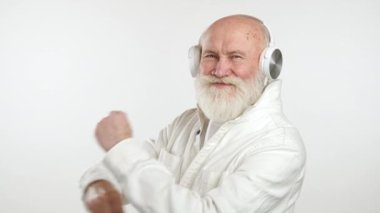 Beyaz tişört ve ceket giyen enerjik yaşlı bir adam, beyaz kulaklıkla müzik dinlerken neşeyle dans ediyor, saf beyaz bir arka plana karşı, canlı enerji ve ritim sergiliyor..