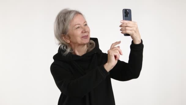一位快乐的老年妇女一边拿着智能手机自私自利 一边享受着科技的发展 她的笑容闪烁着幸福的光芒 穿着休闲的黑色衣服 与现代世界的交往 8K照相机 — 图库视频影像