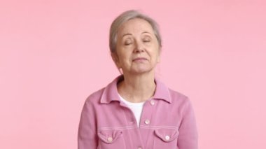 Pembe ceketli olgun bir kadın kararlı bir şekilde kafasını sallayarak güçlü kişisel görüşünü sembolize eden yumuşak pembe bir arka plana karşı jesti ile bir firmaya hayır işareti yapıyor. Kamera 8K HAM. 