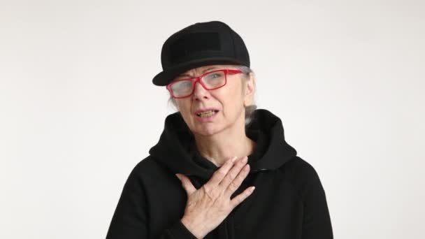 一位身穿黑色服装 头戴帽子的老年妇女在苍白的背景下咳嗽着 紧紧抓住胸部 显得很苦恼 这可能是一种不适或健康问题的迹象 8K照相机 — 图库视频影像