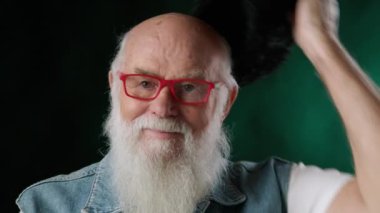 Bu nazik videoda, gür beyaz sakallı ve canlı kırmızı gözlüklü gülümseyen bir büyükbaba yavaşça örgü şapkasını karanlık bir arka plana doğru çıkarırken büyülenir. Kamera 8K HAM. 