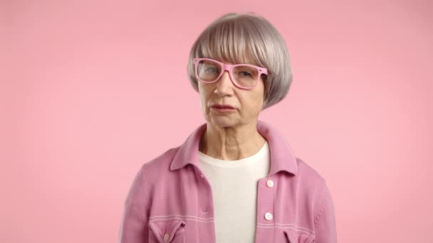 一位留着浅色短发 穿着粉色夹克和眼镜的老太太 在配着粉色背景的时候 没有做手势地摇着头 8K照相机 — 图库视频影像