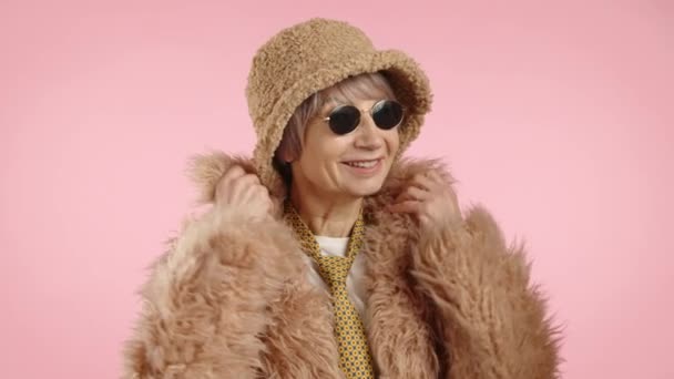 一位时髦的老妇人散发出自信和时尚的气息 穿着时髦的人造毛皮外套 头戴时髦的斗篷帽 戴着圆圆的太阳镜 衬托着柔和的粉色背景 高质量的4K镜头 — 图库视频影像