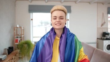 Lezbiyen bir kadın evinde mutluluk saçıyor, elleriyle kalp şekli yaratıyor, LGBT bayrağına sarılı, aşkı ve gururu sembolize ediyor, samimi bir ortamda. Yüksek kalite 4k görüntü