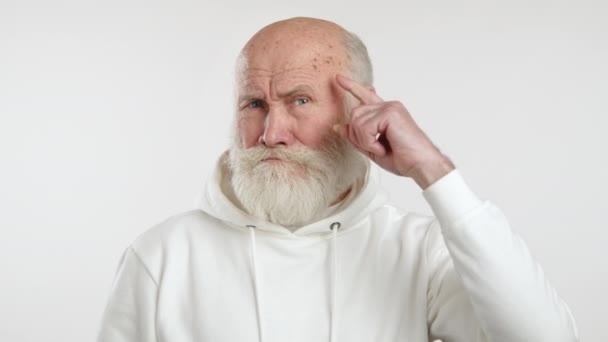 视频中 一个穿着白色帽衫的老人用手指触摸自己的太阳穴 做了一个疯狂的动作 表现出幽默或讽刺的表情 8K照相机 — 图库视频影像
