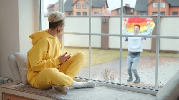 女性が彼女のレズビアンのパートナーが窓の外で遊んで踊るのを見る喜びの瞬間 虹のプライドフラッグを振り 愛と自由のシンボル 高品質の4K映像 — ストック動画