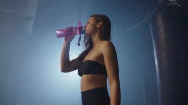 Yoğun bir boks antrenmanından sonra, bir fitness meraklısı susuzluğunu gidermek için durur, su şişesinden bir yudum alarak, sıkı bir spor salonunda su alımının önemini güçlendirir..