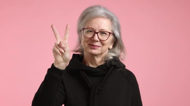 一位戴眼镜 身穿黑色夹克 头戴眼镜的老妇人高兴地做出了一个和平手势 脸上挂着微笑 — 图库视频影像
