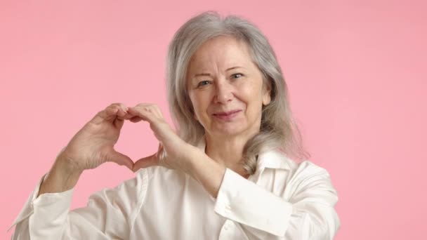 白いシャツの愛情深い高齢女性は 彼女の手でハートシンボルを作り 補完的なパステルピンクの背景に対して暖かさと優しさを示しています カメラ Raw — ストック動画