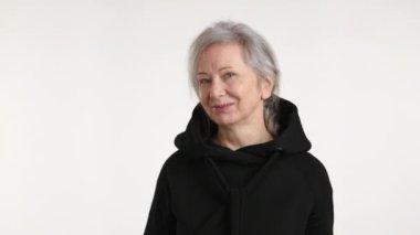 Neşeli, yaşlı bir kadın siyah kapüşonlusunu şakayla ağzına çekiyor, arsız bir sırıtış sergiliyor minimalist beyaz bir arka plana.
