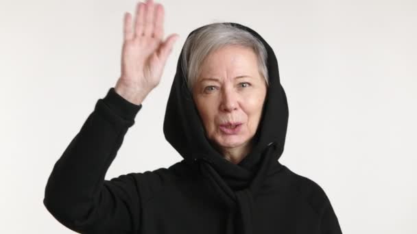 一位穿着休闲黑色衣服的快乐的老年妇女挥挥手愉快地打招呼 与白色背景隔离 象征着温暖和友善 — 图库视频影像