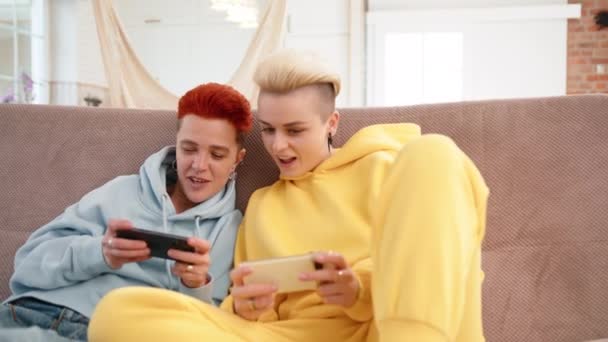 一对穿着舒适休闲装的女同性恋夫妇 在舒适的家庭环境中坐在沙发上 坐在一起笑着 享受着一个竞争激烈的手机游戏 8K照相机 — 图库视频影像