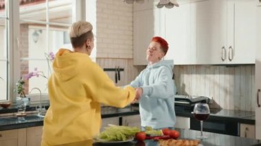 Neşeli lezbiyen bir çift mutfakta neşeli bir anı paylaşıyor, biri güneşli sarı kapüşonlu, diğeri yumuşak mavi, bir bardak şarap ve sıcak atmosfere katılan taze malzemelerle.