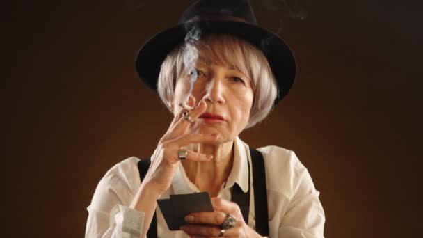 手里拿着一支香烟 一位穿着白衬衫 头戴白帽的快乐的老太太露出了她的扑克牌 在一个舒适的老式环境中 她的脸上闪烁着游戏的兴奋 8K照相机 — 图库视频影像