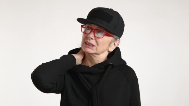 黒いパーカーと帽子を被るスタイリッシュな高齢女性は 首の痛みを表現し 痛みを伴う表現で中立的な背景に立っています 高品質の4K映像 — ストック動画