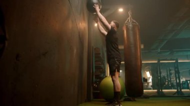 Endüstriyel bir spor salonunun loş ışığında, bir adam bir ilaç topuna tüm vücudunu kullanır, eko yoğun olarak kas ve kardiyovasküler güç oluşturmaya odaklanır.. 