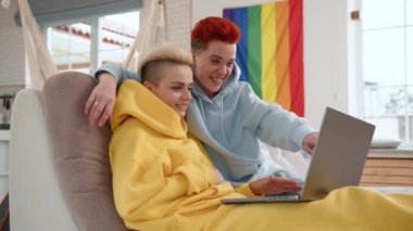 Rahat bir ev ortamında, canlı saç renkleri olan iki genç kadın ortak bir kapüşonluya sarılır, arkalarında asılı duran bir LGBTQ gururunun sembolü olan bir dizüstü bilgisayarla neşeyle meşgul olurlar. Kamera 8K HAM