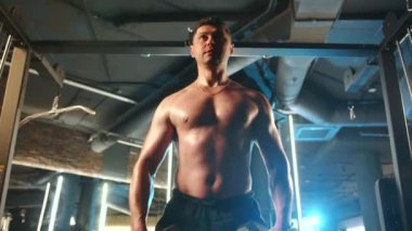 Tişörtsüz ve odaklanmış bir adam modern bir spor salonunda dambıllarıyla yan yana yükselerek omuz kaslarını hedef alıyor, fitness rejimine disiplinli bir yaklaşım sergiliyor.. 