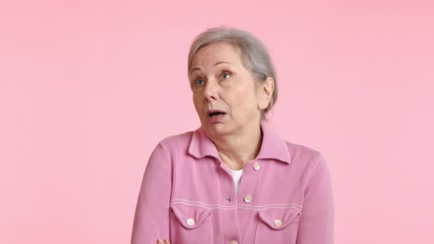 このシーンでは ピンクのジャケットのシニア女性が皮肉な表情を示し 不信感と不透明な状況についての混乱の中で眉を上げ パステルピンクの背景 — ストック動画