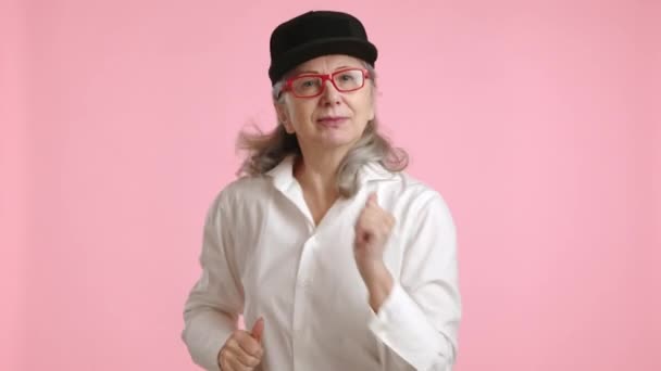 在这段温馨的视频中 一位精力充沛的老妇人快乐地跳着舞 戴着时髦的红色眼镜 头戴时髦的黑色帽子 在柔和的粉色背景下显得更有活力 — 图库视频影像