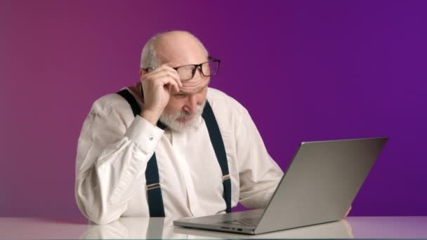 一位恼怒的老人在工作于笔记本电脑时沮丧地摘下了眼镜 这表明他需要在一个充满活力的紫色背景上休息一下 8K照相机 — 图库视频影像