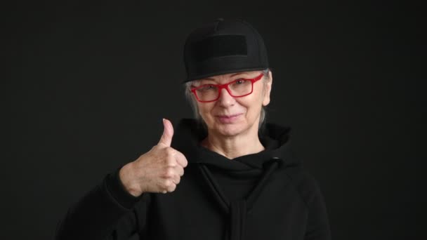 穿着黑色服装 戴着红色眼镜的时髦老年女性竖起大拇指 在深色背景下表现出积极向上的气质 很好的动机内容 8K照相机 — 图库视频影像