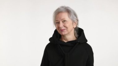 Neşeli, yaşlı bir kadın siyah kapüşonlusunu şakayla ağzına çekiyor, arsız bir sırıtış sergiliyor minimalist beyaz bir arka plana.