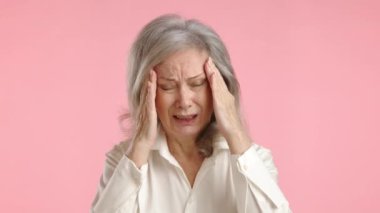 Yetişkin bir iş kadını, günlük eziyetin ortasında stresten kaynaklanan bir baş ağrısı çekerken yüzünü pembe bir arka planda mesleki sorumlulukların yükünü ifade ederek yüzünü buruşturuyor. Kamera 8K HAM. 