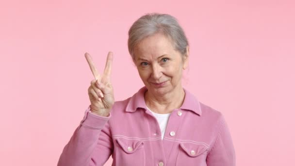 マゼンタのジャケットの高齢女性は 喜んで微笑んでいる間 親指で平和のサインジェスチャーを作るために腕を伸ばしています — ストック動画