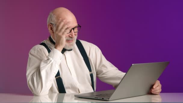 高齢の紳士がラップトップのビデオ通話に没頭し サスペンサーとボウタイで白いシャツをスマートに着てピンクの紫色の背景に設定されています カメラ Raw — ストック動画