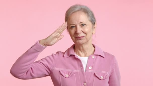 一位泰然自若的老妇人站在那里 双手托着额头敬礼 摆出一种恭敬的军人风度 穿着一件柔软的粉色夹克 衬托着淡淡的粉色背景 — 图库视频影像