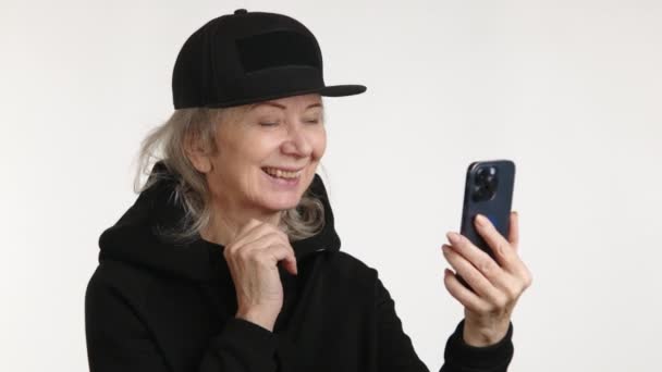 一位老妇人一边享受着视频通话的乐趣 一边戴着时髦的黑色帽子 穿着朴素的白色背景的衣服 从中散发出欢乐的光芒 — 图库视频影像