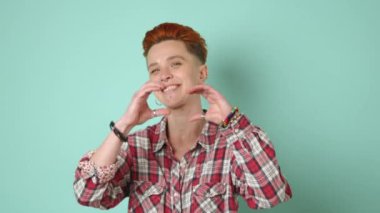 Lezbiyen bir kadın elleriyle kalp şekli oluşturur. Parlak bir gülümseme ile sevgi ve pozitiflik iletir. Yumuşak bir deniz mavisi arkaplanının önünde durur. Kamera 8K HAM. 