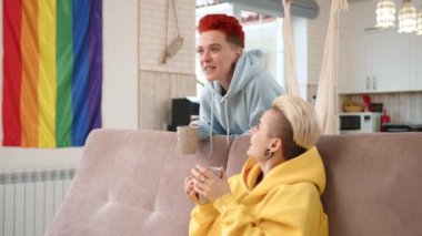 Evlerinin sıcaklığında, lezbiyen bir çift rahat bir çay molası veriyor ve arka planda LGBT bayrağının rahatlatıcı varlığıyla sohbet ediyorlar. Yüksek kalite 4k görüntü