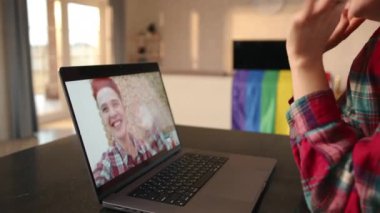 Bir lezbiyen, partnerini bir video çağrısında karşılarken heyecanını ve sevgisini ifade eder, onun samimi gülümsemesi dijital alanda derin bir bağlantı kurar, arka planda bir LGBT bayrağı ile.