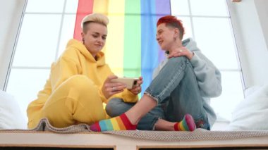 Neşeli lezbiyen bir çift, rahat bir şekilde pencerenin kenarında oturur, birlikte telefona bakarken kahkahalarla güler, sıcak ve sevgi dolu bir bağ sergilerler. Kamera 8K HAM.