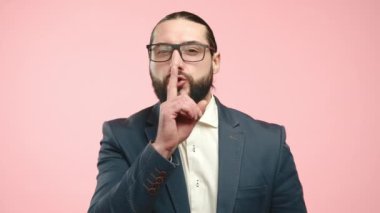 Zarif bir göz giysisi ve özel dikim takım elbiseli tatlı bir işadamı parmaklarını dudaklarına koyar, sessiz bir şekilde, yumuşak pembe bir arka plana doğru, gizli bir iletişim hissi uyandırır. Kamera 8K HAM