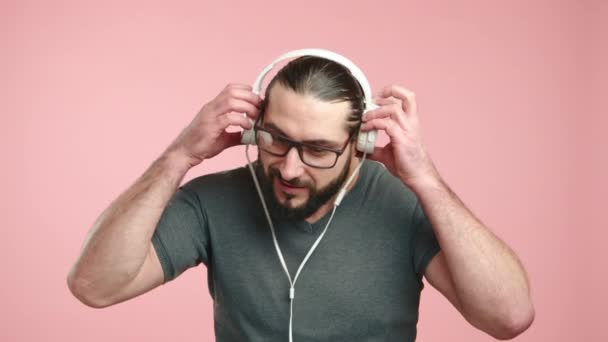 カジュアルなTシャツの男がヘッドフォンを着て コンテンツに目を閉じ 明らかに音楽と瞬間を楽しんで 快適なピンクの背景に設定しました カメラ Raw — ストック動画