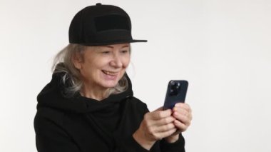Neşeli, yaşlı bir kadın, telefonuna bir mesaj yazarken gülümserken görülüyor. Siyah bir şapkası ve temiz beyaz bir geçmişi olan bir giysisi var.. 