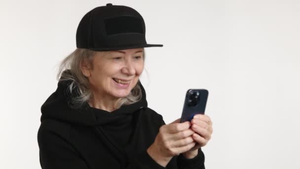 一位快乐的老妇人一边在手机上留言 一边微笑着 戴着一顶随意的黑色帽子 穿着洁白的衣服 — 图库视频影像