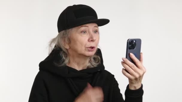 一位年长的女性在使用智能手机进行视频通话时表现出失望的迹象 她身穿黑色的帽子和高领毛衣 背景是简约的白色 — 图库视频影像