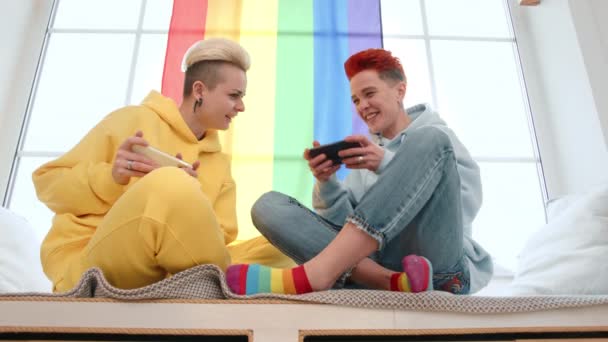 在同性恋关系中 两位可爱的年轻女性在智能手机上玩游戏 她们蜷缩在窗前 打着Lgbtq骄傲的旗子 8K照相机 — 图库视频影像