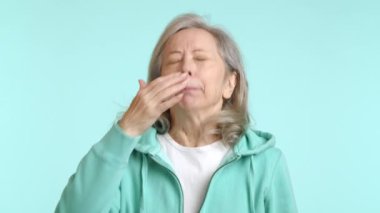 Yaşlı kadın eline hapşırır, doğal hapşırma refleksi gösterir, ferah, açık mavi bir arka plana karşı sıradan bir nane yeşili kapüşon giyer. Kamera 8K HAM. 