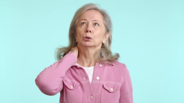 ピンクの服を着た高齢女性は 不快感を抱き 痛みを意味するために首を握り 医療と高齢者の健康を強調した落ち着いた青色の背景に設定しました カメラ Raw — ストック動画