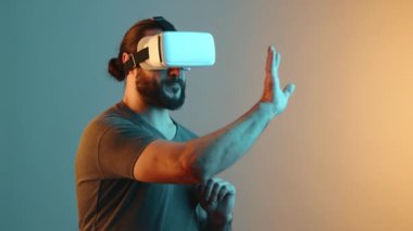 Bir adam sanal gerçeklik deneyimine kendini tamamen kaptırmış durumda, sanal gerçeklik gözlüğü kullanarak dijital dünyayla etkileşime giriyor, elini kaldırıp jest yapıyor, sıcak renkli bir arkaplanda. Kamera 8K HAM. 